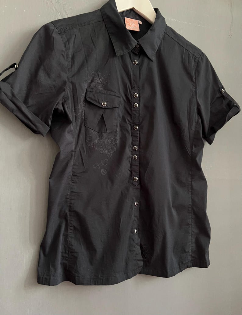 May include: A black short-sleeved button-down shirt with a collared neckline. The shirt features a chest pocket on the left side and decorative detailing. The sleeves can be rolled up and secured with a button. The shirt is hanging on a white hanger.