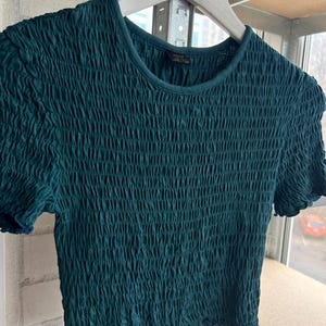 K&ouml;nnte beinhalten: Ein teal-gr&uuml;nes, kurz&auml;rmeliges Crop-Top mit einem strukturierten, gerafften Design. Das Oberteil h&auml;ngt an einem wei&szlig;en Holzkleiderb&uuml;gel. Der Halsausschnitt und die &Auml;rmel haben eine dezente, dunklere Einfassung, und der Stoff scheint ein weiches, dehnbares Material zu sein.