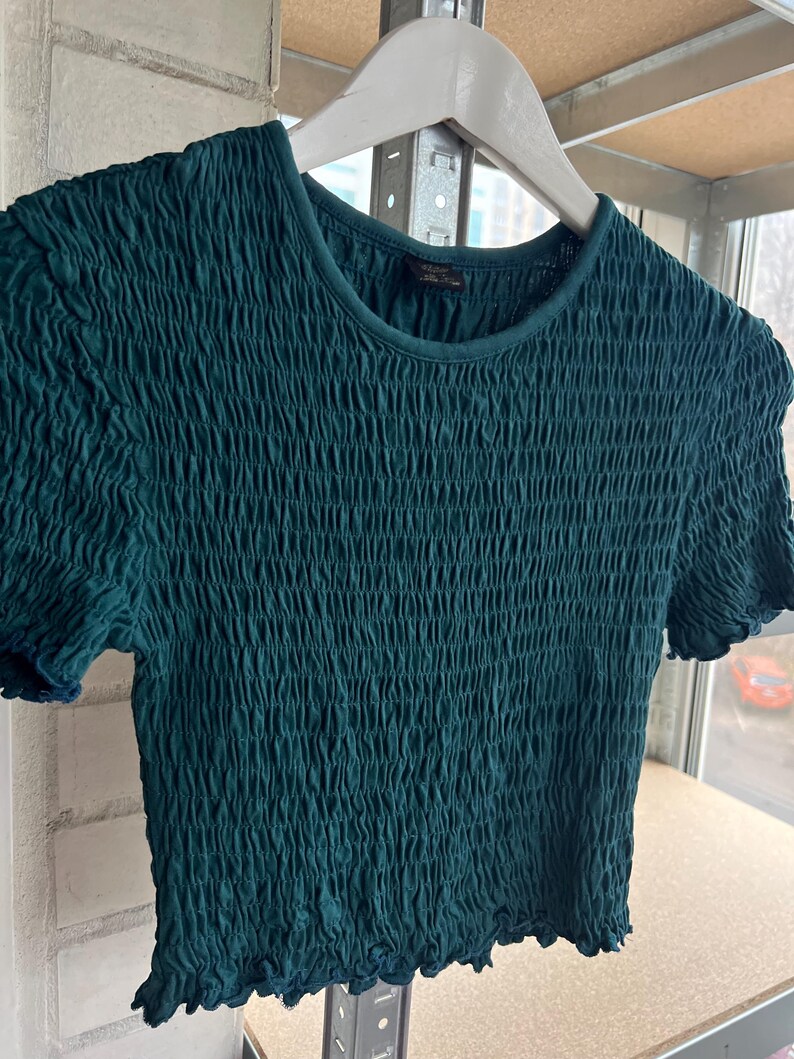 K&ouml;nnte beinhalten: Ein teal-gr&uuml;nes, kurz&auml;rmeliges Crop-Top mit einem strukturierten, gerafften Design. Das Oberteil h&auml;ngt an einem wei&szlig;en Holzkleiderb&uuml;gel. Der Halsausschnitt und die &Auml;rmel haben eine dezente, dunklere Einfassung, und der Stoff scheint ein weiches, dehnbares Material zu sein.