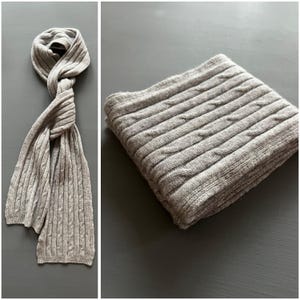 May include: A gray cable knit scarf with a textured pattern. The scarf is folded in half and laid flat on a gray surface.
