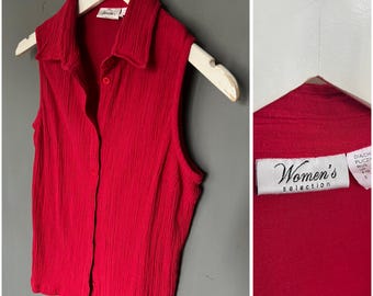 May include: A sleeveless red collared shirt with a button-down front, hanging on a white wooden hanger. The shirt has a textured, ribbed appearance. The tag reads "Women's selection".