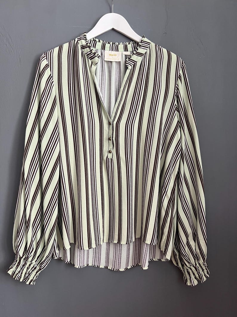 May include: A green and black striped long-sleeved blouse with a button closure and a ruffled collar. The blouse has a slightly gathered hemline and elastic cuffs.