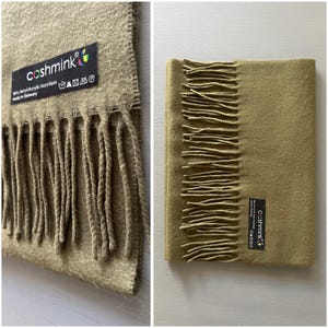 May include: Olive green scarf with fringe detail. The scarf has a rectangular shape and features a label that reads "cashmink". The scarf is made of 85% Acrylic and 15% Polyamide. The scarf is a solid color and has a soft texture.