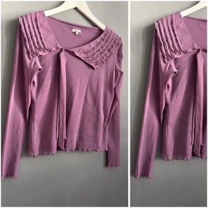 Könnte beinhalten: Ein langärmliges, malvenfarbenes Oberteil mit einem einzigartigen Kragendesign. Der Kragen hat plissierten Stoff und eine Krawatte. Das Oberteil hat einen gewellten Saum und wird auf einem weißen Kleiderbügel vor grauem Hintergrund präsentiert.