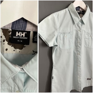 Puede incluir: Una camisa de manga corta de color azul claro con botones, dos bolsillos en el pecho y el logotipo de Helly Hansen en el bolsillo izquierdo y el dobladillo inferior derecho. La camisa tiene un cuello abotonado blanco y una tapeta de botones blanca.