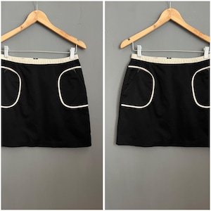 Könnte beinhalten: Schwarzer Minirock mit weißen Paspeldetails und Taschen. Der Rock hat ein schlichtes, klassisches Design.