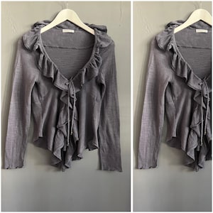 May include: A gray long-sleeved cardigan with a ruffled trim and a tie closure. The cardigan is hanging on a white hanger. The fabric appears to be lightweight and has a subtle texture.