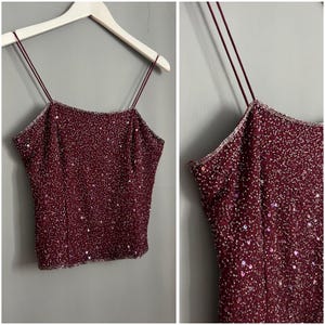 Puede incluir: Un top de tirantes de color burdeos con lentejuelas. El top está hecho de una tela suave y elástica y tiene un aspecto delicado y femenino.