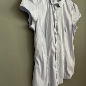 Op de afbeelding: Lichtblauw overhemd met korte mouwen en een wit stippenpatroon. Het overhemd heeft een ronde kraag en hangt aan een witte hanger. Het overhemd is gemaakt van een lichte stof.