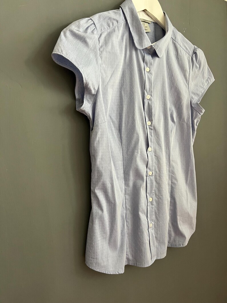 Op de afbeelding: Lichtblauw overhemd met korte mouwen en een wit stippenpatroon. Het overhemd heeft een ronde kraag en hangt aan een witte hanger. Het overhemd is gemaakt van een lichte stof.