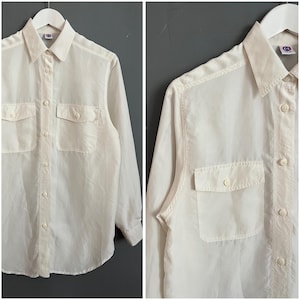 Pode incluir: Uma camisa branca de manga comprida com botões e dois bolsos no peito. A camisa tem uma gola clássica e um caimento ligeiramente solto.