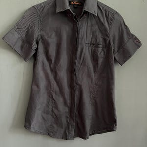 May include: Dark grey short-sleeved shirt with vertical pinstripes. The shirt has a small chest pocket and is hanging on a white hanger. The shirt has a collar and the sleeves are cuffed.