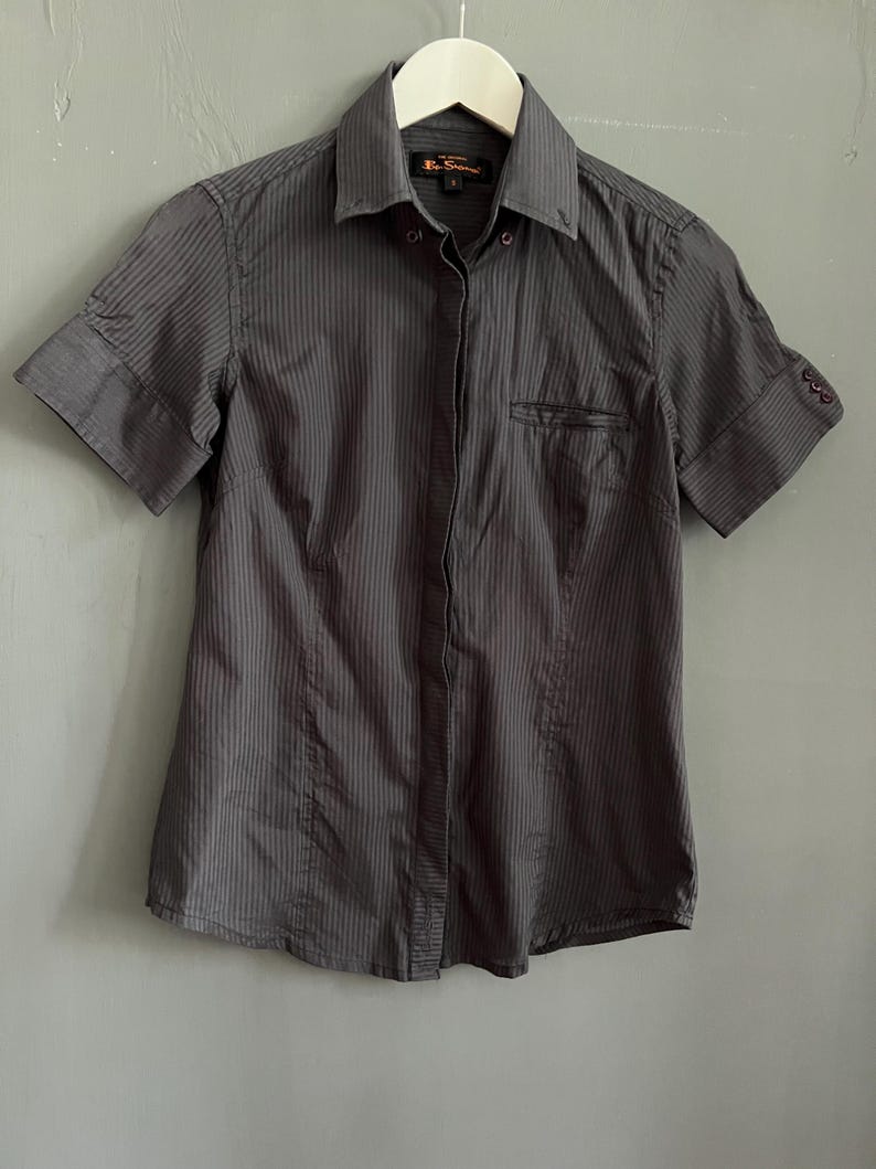 May include: Dark grey short-sleeved shirt with vertical pinstripes. The shirt has a small chest pocket and is hanging on a white hanger. The shirt has a collar and the sleeves are cuffed.