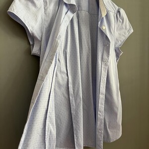 Op de afbeelding: Een lichtblauwe blouse met korte mouwen, een witte kraag en kleine stippen. De blouse is opengeknoopt en onthult de binnenstof. Het kledingstuk hangt aan een witte hanger tegen een grijze achtergrond.