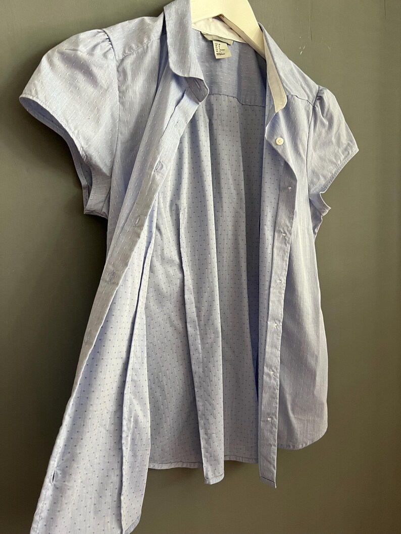 Op de afbeelding: Een lichtblauwe blouse met korte mouwen, een witte kraag en kleine stippen. De blouse is opengeknoopt en onthult de binnenstof. Het kledingstuk hangt aan een witte hanger tegen een grijze achtergrond.
