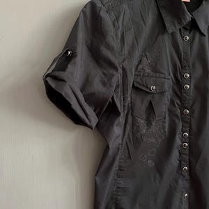 May include: Black short-sleeve button-up shirt with two chest pockets and a collared neckline. The shirt has a roll-tab sleeve design and decorative embroidery. The shirt is made of a lightweight fabric.