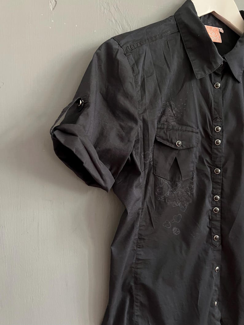 May include: Black short-sleeve button-up shirt with two chest pockets and a collared neckline. The shirt has a roll-tab sleeve design and decorative embroidery. The shirt is made of a lightweight fabric.