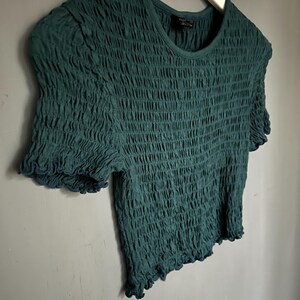 K&ouml;nnte beinhalten: Ein teal-gr&uuml;nes Oberteil mit kurzen &Auml;rmeln und einem strukturierten, ger&uuml;schten Design. Das Shirt hat einen runden Ausschnitt und ger&uuml;schte Kanten an den &Auml;rmeln und am Saum. Das Kleidungsst&uuml;ck h&auml;ngt an einem wei&szlig;en Kleiderb&uuml;gel vor einer grauen Wand.