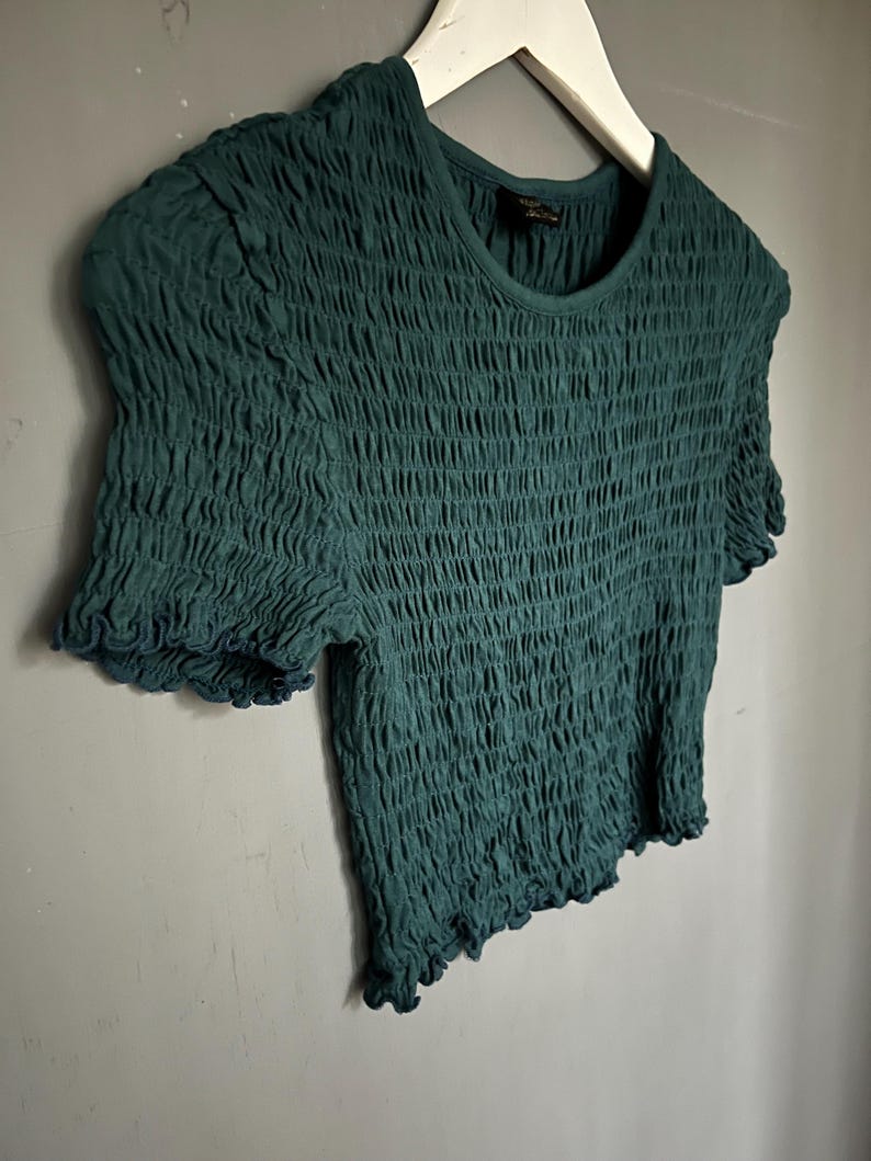K&ouml;nnte beinhalten: Ein teal-gr&uuml;nes Oberteil mit kurzen &Auml;rmeln und einem strukturierten, ger&uuml;schten Design. Das Shirt hat einen runden Ausschnitt und ger&uuml;schte Kanten an den &Auml;rmeln und am Saum. Das Kleidungsst&uuml;ck h&auml;ngt an einem wei&szlig;en Kleiderb&uuml;gel vor einer grauen Wand.