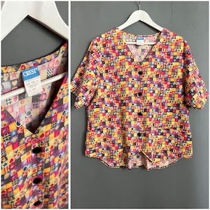 Puede incluir: Una camisa de manga corta con botones y un estampado geométrico colorido con corazones, estrellas y cuadrados en tonos amarillos, rojos, morados y azules. La camisa tiene cuello en V y cierre de botones en la parte delantera. La etiqueta dice "CREST" con el número de estilo "1108" y la talla "2XL".