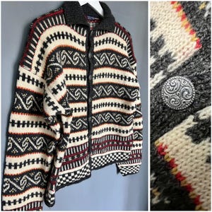 Könnte beinhalten: Knopf-Cardigan mit geometrischem Muster in Creme, Schwarz und Rot. Der Pullover hat einen dunkelgrauen Kragen und Manschetten sowie dekorative silberne Knöpfe. Das Design umfasst horizontale Streifen und stilisierte Formen.