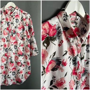 May include: A white button-down shirt with a floral print in shades of pink and gray. The shirt has a long sleeve and a collared neckline.