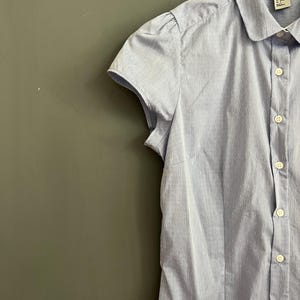 Op de afbeelding: Een lichtblauw overhemd met korte mouwen en een subtiel stippenpatroon. Het overhemd heeft een ronde kraag en kleine, gepofte mouwen. Het kledingstuk hangt aan een witte hanger tegen een gedempte groene achtergrond.