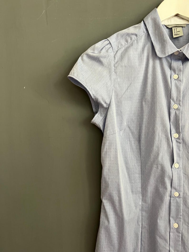 Op de afbeelding: Een lichtblauw overhemd met korte mouwen en een subtiel stippenpatroon. Het overhemd heeft een ronde kraag en kleine, gepofte mouwen. Het kledingstuk hangt aan een witte hanger tegen een gedempte groene achtergrond.