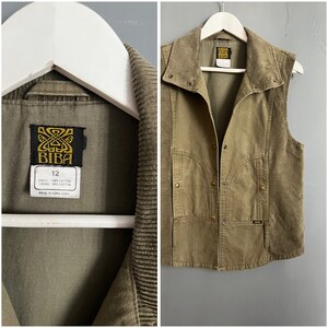 Op de afbeelding: Een groene corduroy vest met een drukknoopsluiting en twee zakken. De vest heeft een label met de tekst "BIBa" en "12". De vest is gemaakt in Hong Kong.