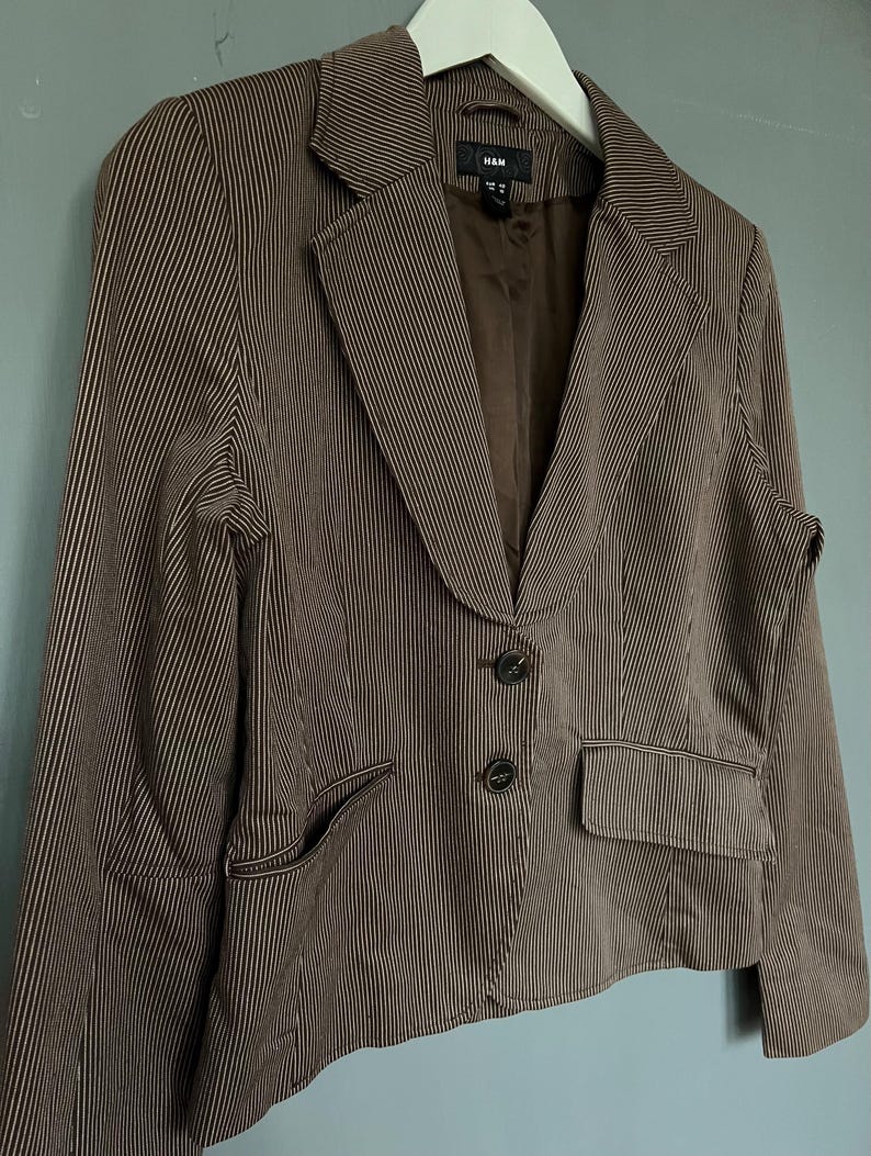 K&ouml;nnte beinhalten: Ein brauner Blazer mit wei&szlig;en Streifen, zwei Kn&ouml;pfen und einer Tasche auf der linken Seite. Der Blazer hat ein Etikett mit der Aufschrift "H&M".