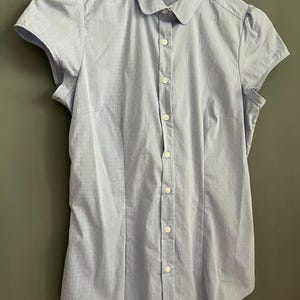 Op de afbeelding: Een lichtblauw overhemd met korte mouwen en een kleine, ronde kraag. Het overhemd heeft een subtiel streepjespatroon en kleine, gepofte mouwen. Het overhemd hangt aan een witte hanger.