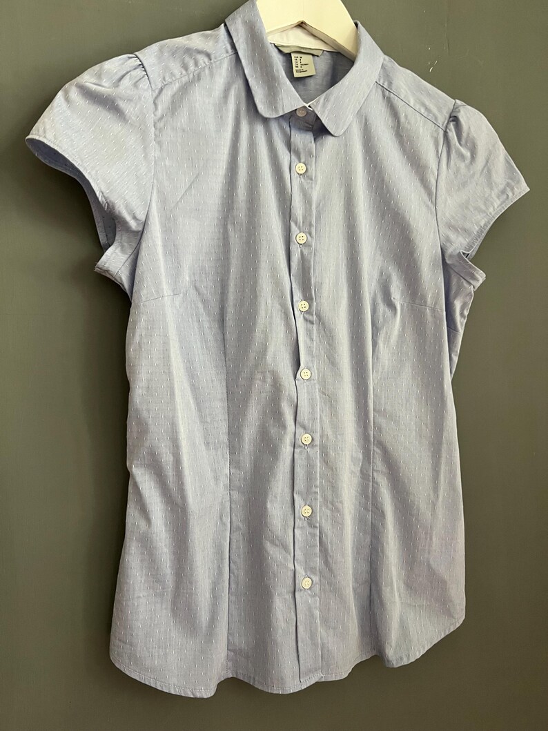 Op de afbeelding: Een lichtblauw overhemd met korte mouwen en een kleine, ronde kraag. Het overhemd heeft een subtiel streepjespatroon en kleine, gepofte mouwen. Het overhemd hangt aan een witte hanger.