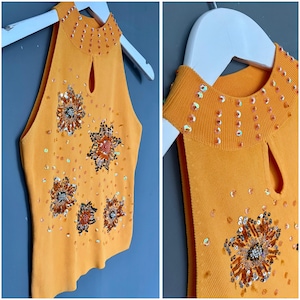 May include: Orange halter top with a keyhole neckline and embellished with sequins and beaded floral designs. The top is sleeveless and has a ribbed texture. The collar and floral designs are adorned with sequins. The top is hanging on a white hanger.