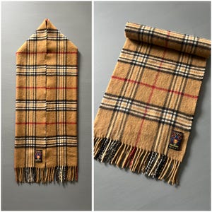 May include: A tan plaid scarf with black, red, and white lines. The scarf has a fringed edge and a small label with text and a logo. The scarf is displayed flat and rolled up.