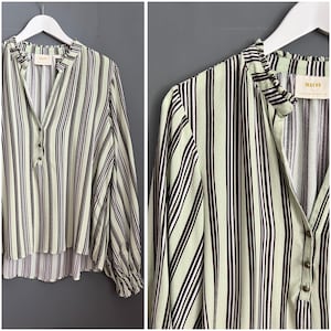 May include: A green and brown striped long-sleeved shirt with a ruffled collar and a button closure. The shirt has a relaxed fit and a slightly longer back hem. The brand name "MAEVA" is printed on the back of the shirt.
