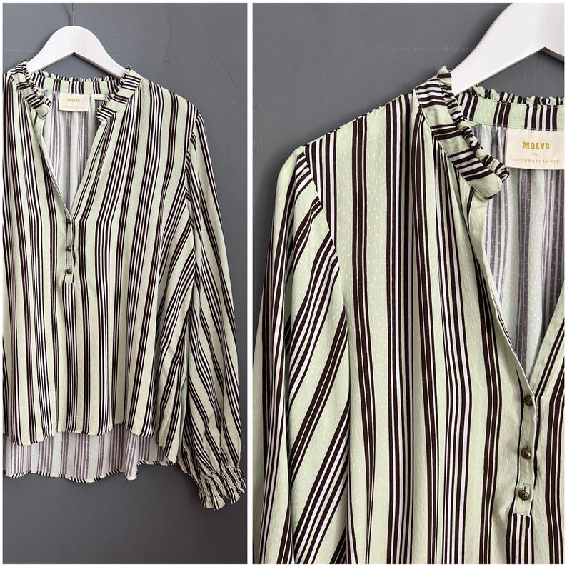 May include: A green and brown striped long-sleeved shirt with a ruffled collar and a button closure. The shirt has a relaxed fit and a slightly longer back hem. The brand name "MAEVA" is printed on the back of the shirt.