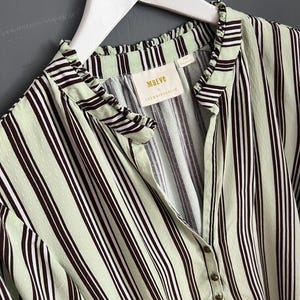 May include: A green and black striped shirt with a ruffled collar and a button closure. The shirt has a small label that reads "Maeve by Anthropologie".