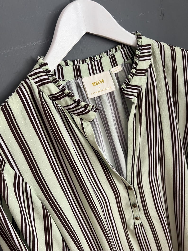 May include: A green and black striped shirt with a ruffled collar and a button closure. The shirt has a small label that reads "Maeve by Anthropologie".