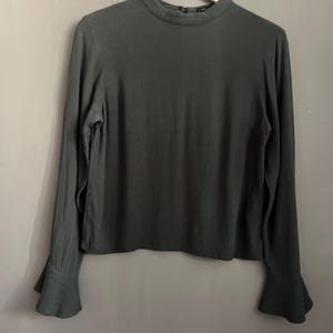 May include: A dark green long-sleeved blouse with a high neckline and flared sleeves. The blouse is hanging on a wooden hanger against a neutral gray background. The fabric appears to be a soft, flowing material.