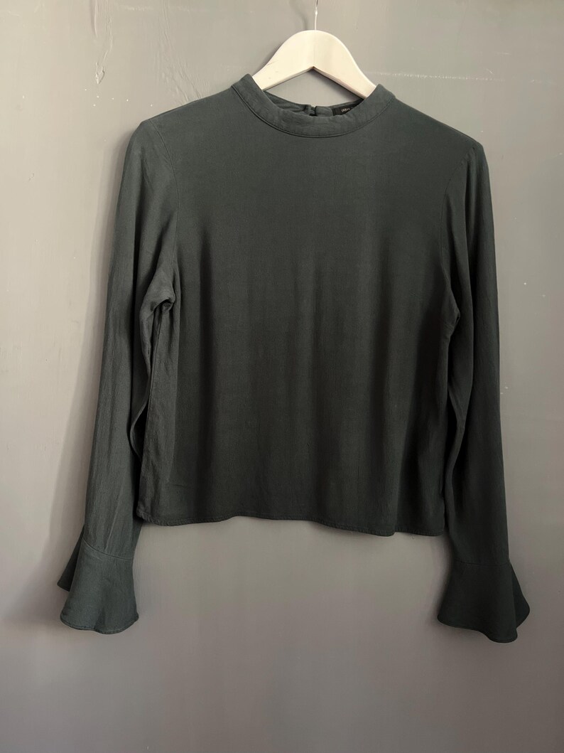 May include: A dark green long-sleeved blouse with a high neckline and flared sleeves. The blouse is hanging on a wooden hanger against a neutral gray background. The fabric appears to be a soft, flowing material.