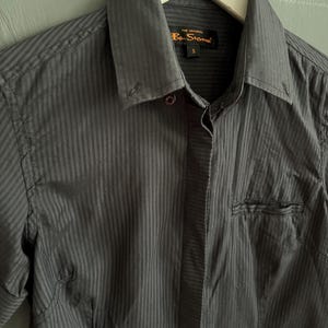 May include: A dark grey striped button-down shirt. The shirt features a pointed collar, a chest pocket, and the brand name "Ben Sherman" is visible on a tag. The shirt is hanging on a white hanger.