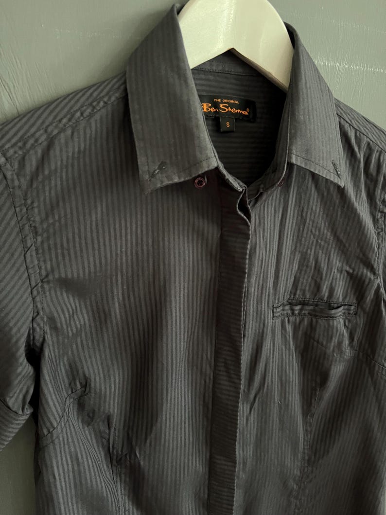 May include: A dark grey striped button-down shirt. The shirt features a pointed collar, a chest pocket, and the brand name "Ben Sherman" is visible on a tag. The shirt is hanging on a white hanger.