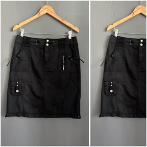 Könnte beinhalten: Schwarzer Cargo-Rock mit mehreren Taschen, einem Reißverschluss und dekorativen Kordelzügen. Der Rock hängt an einem Holzbügel vor grauem Hintergrund. Der Rock hat einen knielangen Saum.