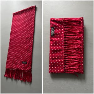 May include: A red scarf with a white polka dot pattern and fringe. The scarf has a label that reads "TOOTAL SCARF". The scarf is made of a soft material and is likely for fashion or warmth.