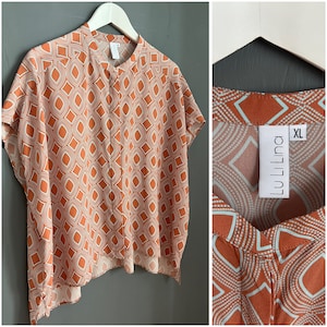 Puede incluir: Una blusa holgada de manga corta, con estampado geométrico naranja y blanco, cuello mandarín y cierre de botón. La blusa está hecha de una tela ligera y tiene un corte holgado. La etiqueta de la blusa dice "LuLiLina" y "XL".
