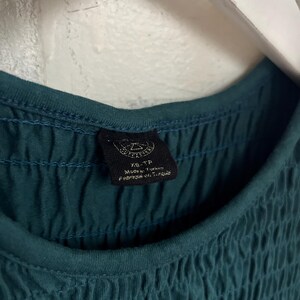K&ouml;nnte beinhalten: Nahaufnahme eines teal-farbenen Oberteils mit einem strukturierten, ger&uuml;schten Oberteil. Der Ausschnitt ist ein einfaches, abgerundetes Design. Ein schwarzes Etikett ist sichtbar, mit dem Text "XS-TP Made in Turkey Fabriqu&eacute; en Turquie."
