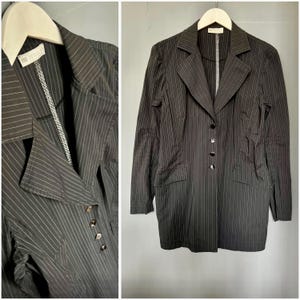 May include: Black pinstripe blazer with a notched collar, three-button closure, and two front pockets. The blazer is hanging on a wooden hanger. The lining is white with a textured design.