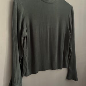 May include: A long-sleeved, dark green blouse with a high neckline and flared sleeves. The blouse is hanging on a wooden hanger against a neutral gray background. The fabric appears to be a soft, flowing material.