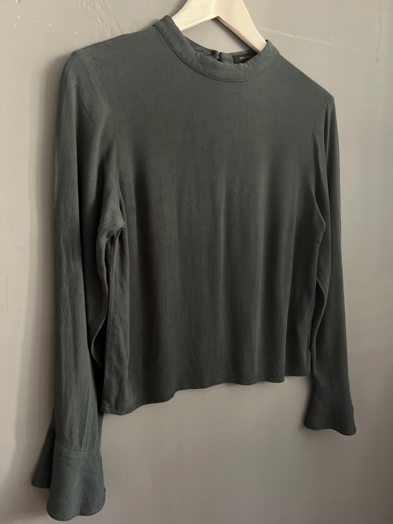 May include: A long-sleeved, dark green blouse with a high neckline and flared sleeves. The blouse is hanging on a wooden hanger against a neutral gray background. The fabric appears to be a soft, flowing material.