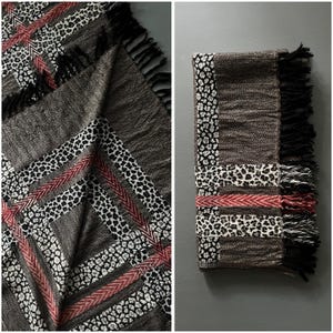 May include: A folded, patterned blanket with a mix of textures and colors. The design includes sections of gray, black, white, and red. The blanket has a fringed edge and a geometric pattern with animal print accents.
